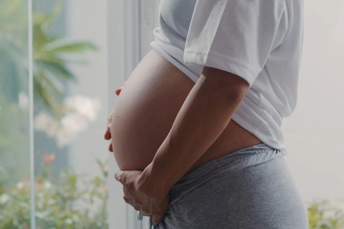 Foto colorida de mulher grávida com a mão na barriga em vrente a uma estrutura de vidro - Metrópoles
