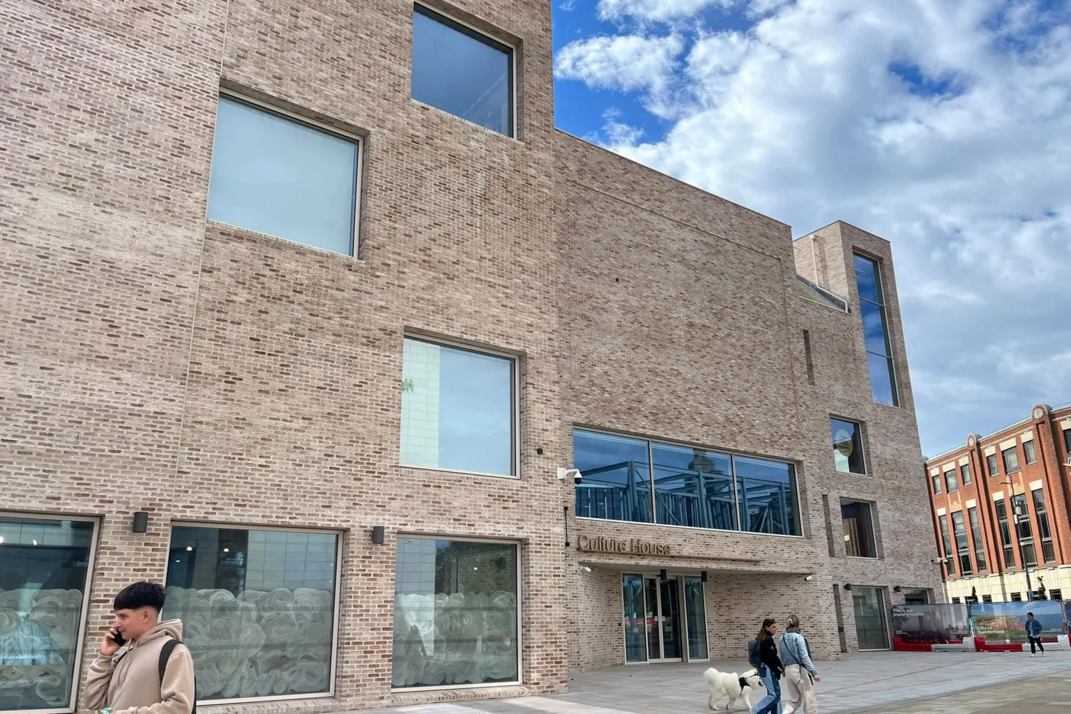 BBC / Kay Davidson Culture House is a multi-storey beige brick building dotted with variously shaped and sized windows. Above the main entrance are the words "Culture House". Several people are walking across the paved square in front of the building.