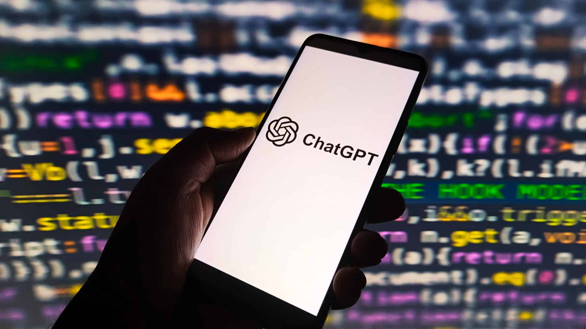 Ao fundo, tela com códigos de programação; à frente, mão de uma pessoa segurando um celular que exibe o logo do ChatGPT