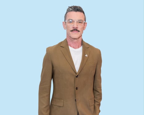 Luke Evans with a quiff, moustache and wearing transparent glasses and a brown suit with white T-shirt
